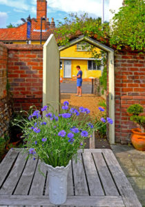 Image of blue cornflowes in white vase on table loooking through open gate in red brick wall out to The Street beyond
