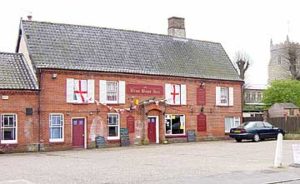 Large and long red bricked building with large flat straight tiled roof and one chimney with a car park in front and the church just visible on the right. A number of signs are on and leaning against the building the main sign being the pub’s dark read sign saying “Blue Boar Inn”.