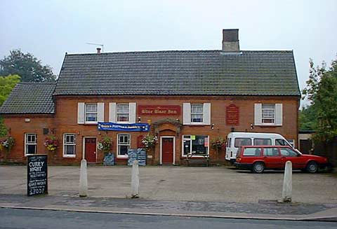 Fairly recent photograph of the Blue Boar taken from the other side of the road. Large and long red bricked building with large flat straight tiled roof. A couple of vehicles in the car park and a Curry Night sign on the edge of the car park. A number of other signs and a banner are on the building itself as well as the pub’s dark read sign saying “Blue Boar Inn”.
