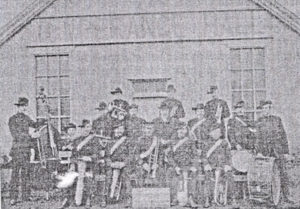 An old rather fuzzy black and white photograph showing a group of men, including Frank Nunn with a double bass, Wilfred Nunn and Harry Nunn, outside the Temperance Hall, all are wearing hats and they are holding various musical instruments.