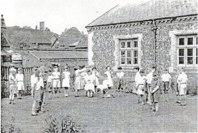 An old black and white photograph of children dressed in white clothing playing in front of Walsham School shortly before it closed.