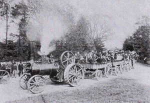 A black and white old photograph of a steam engine pulling a trailer holding a lot of people.